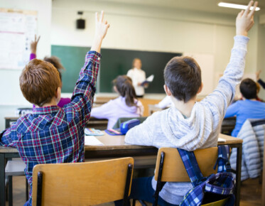 Schoolchildren at classroom with raised hands answering teacher's question.