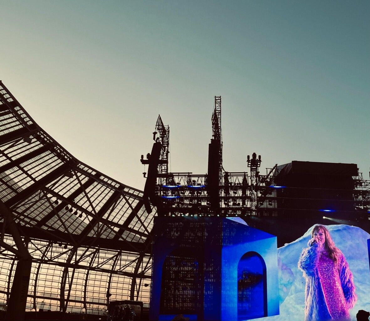 Taylor Swift seisomassa stadionin konserttilavalla mikrofoni kädessä auringonlaskun aikaan, valokuva
