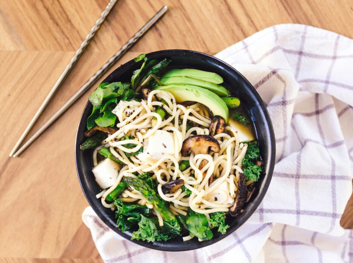 Ramen kulho puisella pöydällä. Kuvassa näkyy myös syömäpuikot ja valkoharmaa keittiöpyyde.