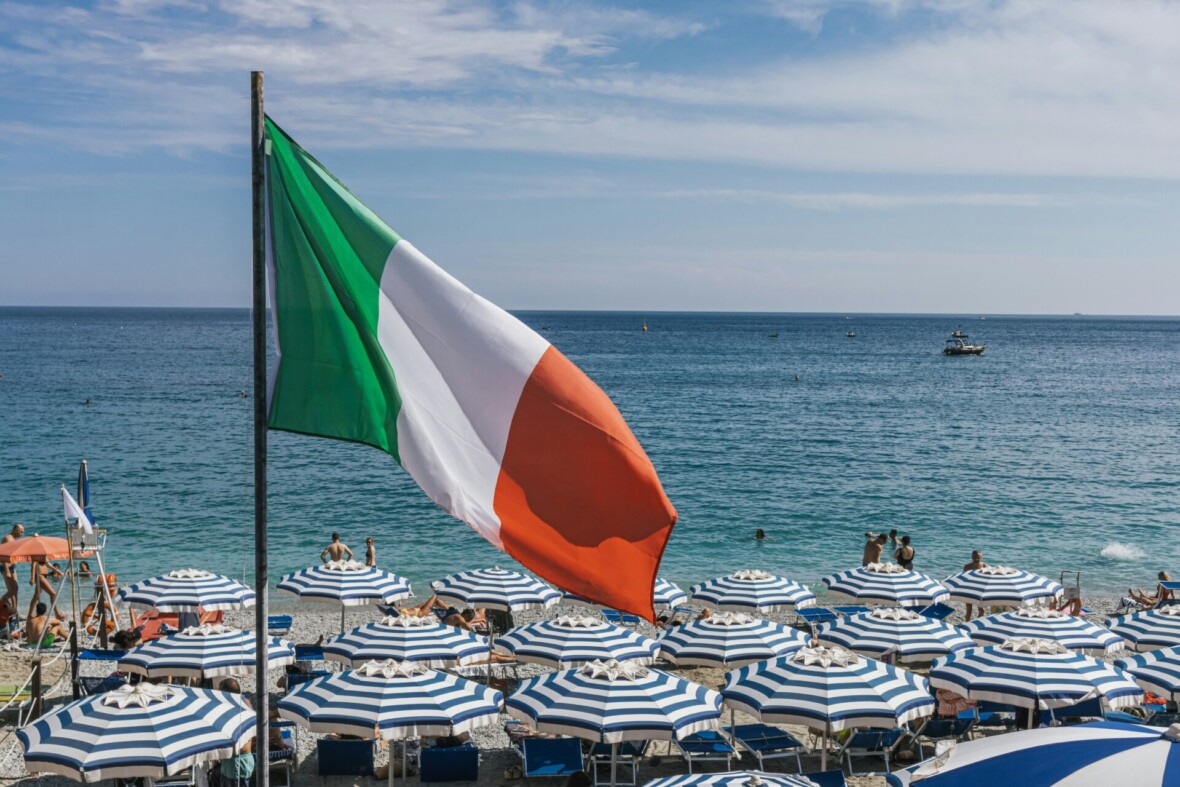 Italian lippu rannalla. Taustalla turkoosi meri ja raidallisia aurinkovarjoja