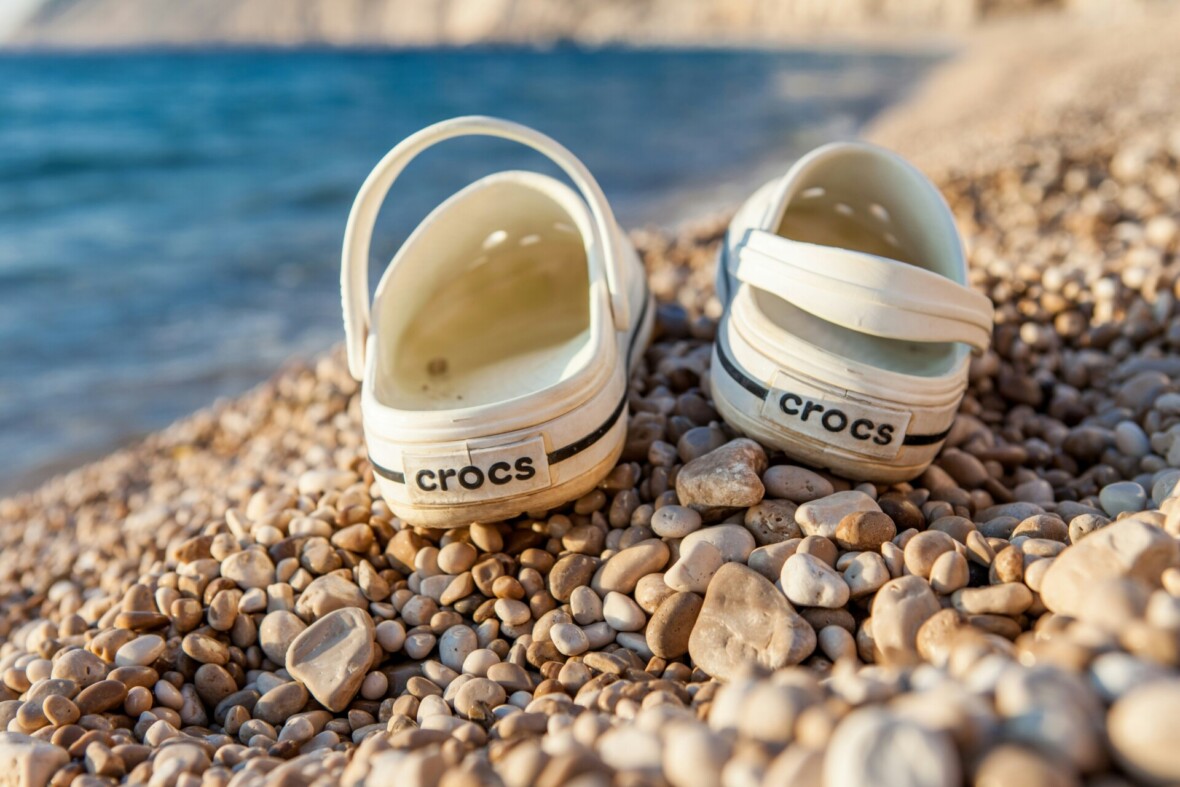 Valkoiset Crocs-kengät kuvattuna takaapäin vaalealla kivirannalla. Vesiraja näkyy taustalla.