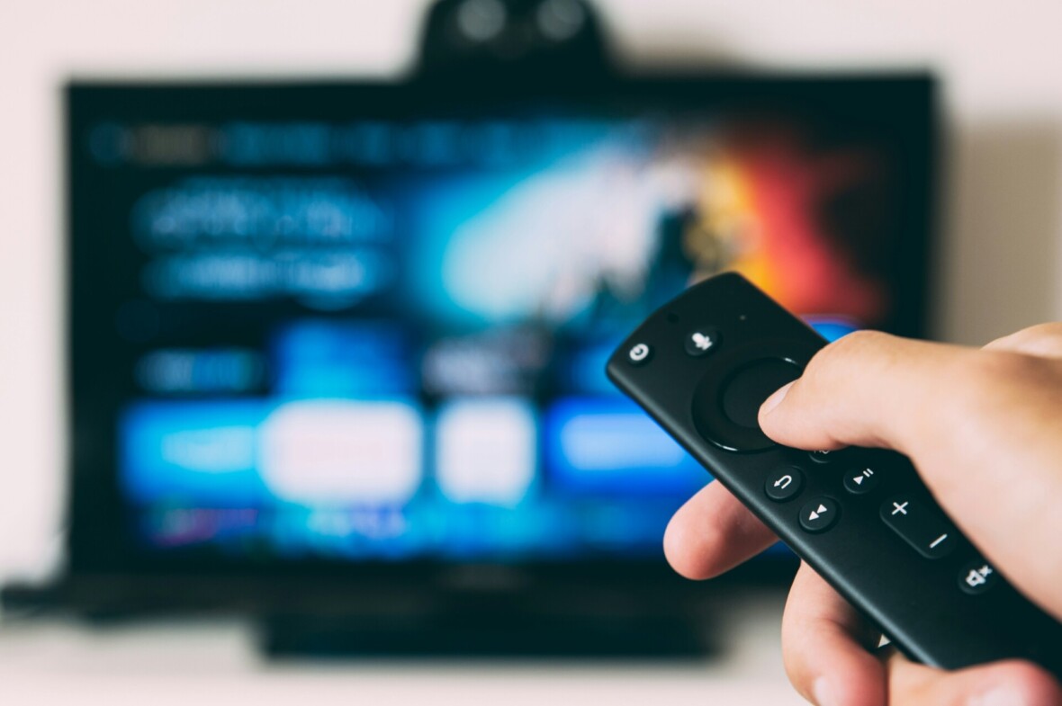 Kuvassa on käsi joka pitelee television kaukosäädintä. Taustalla näkyy sumennettuna televisio, jossa on auki suoratoistopalvelun etusivu.